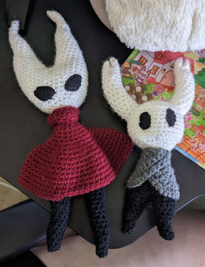 2 crochet plushies of the characters Hornet & Hollow Knight from the video game Hollow Knight. Hornet is made from white, black, and maroon yarn. Hollow Knight is made out of white, black, and gray yarn.