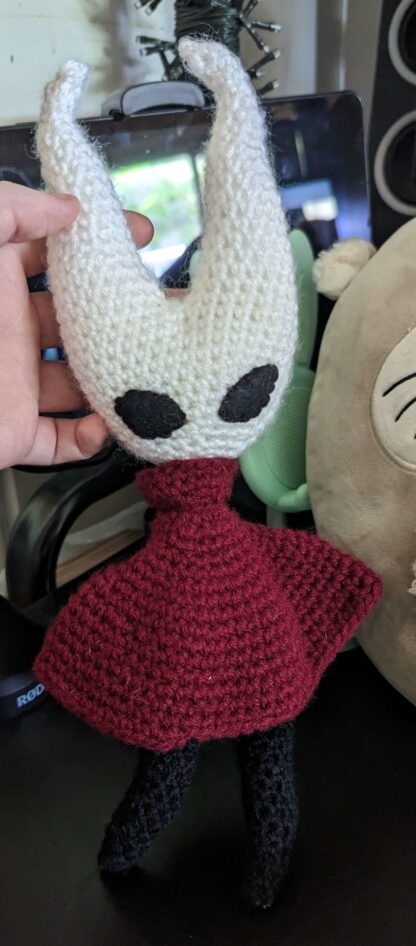 A crochet plushie of the character Hornet from the video game Hollow Knight. The plushie is made from white, black, and maroon yarn.