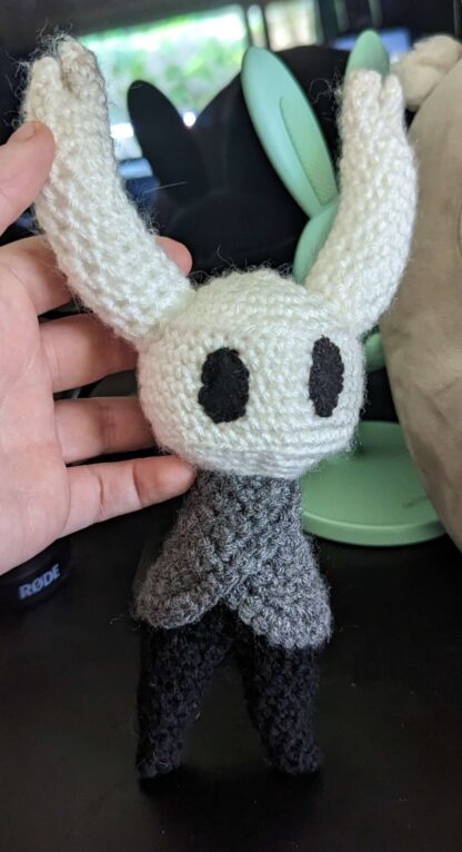 A crochet plushie of the character Hollow Knight from the video game Hollow Knight. The plushie is made out of white, black, and gray yarn.